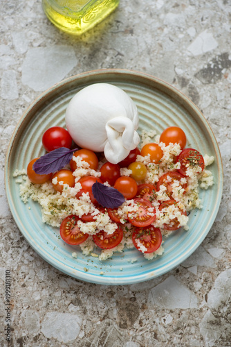 Burrata cheese with cherry tomatoes and couscous on a turquoise plate, vertical shot on a light-brown granite background, elevated view