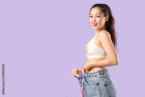 Photos Young Asian woman in loose jeans measuring waist on lilac background