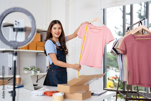 Asian beautiful female bloggers smiling with happiness and joyfulness, greeting people, recording streaming video clip online selling clothes using lighting, laptop, tablet in indoor studio