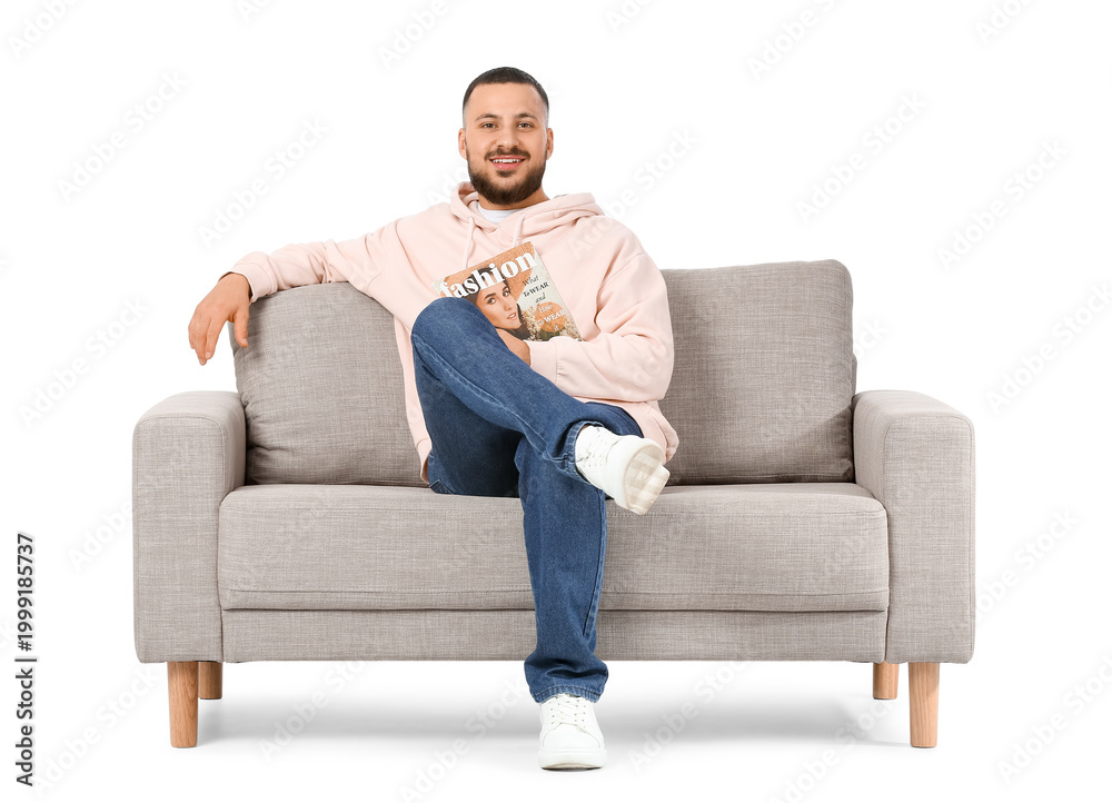 Fototapeta premium Young man with magazine sitting on grey sofa against white background