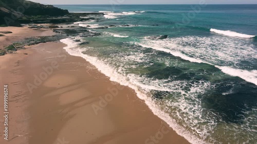 Aerial shot of the ocean with waves doing swirls and patters, rocks, sand and beach