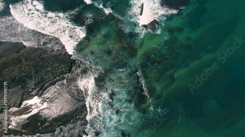 Aerial shot of the ocean with waves doing swirls and patters, rocks, sand and beach