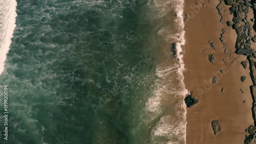 Aerial shot of the ocean with waves doing swirls and patters, rocks, sand and beach