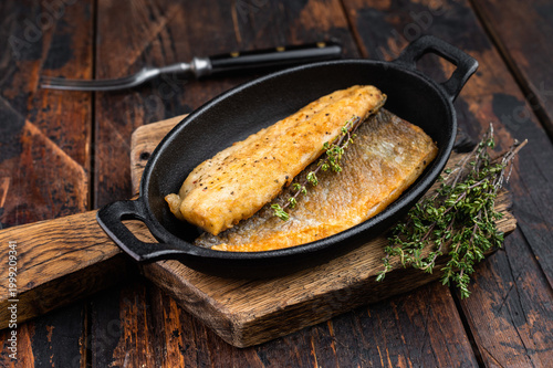 Pan fried seabass or sea bass fillet served in rustic skillet with herbs on wooden table, cooked branzino seafood meal photographed in overhead restaurant style.