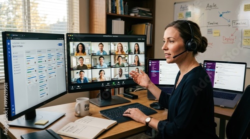 A woman wearing a headset and headphones is engaged in a video conference call, pointing at a computer screen displaying a Microsoft Teams interface with multiple participants.