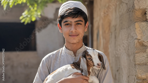 A young boy in traditional clothing holds a small goat, smiling at the camera in a sunlit outdoor setting.