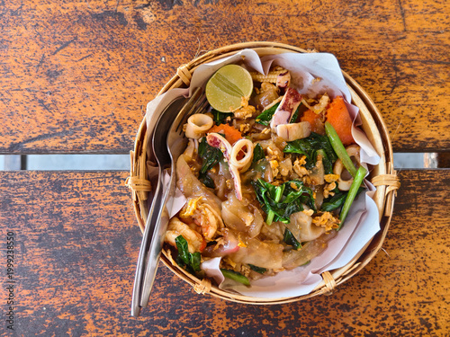 Authentic Thai Pad See Ew. Stir-fried wide rice noodles with shrimp, squid, and kale in sweet soy sauce. Beautifully served in a rustic bamboo basket on a wooden table. High-quality food photography.