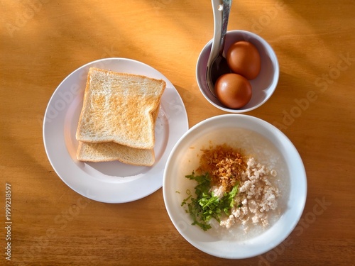 Top view of a healthy Asian breakfast set: savory rice congee with minced pork and ginger, served with crispy toast and two boiled eggs on a wooden table with morning sunlight.