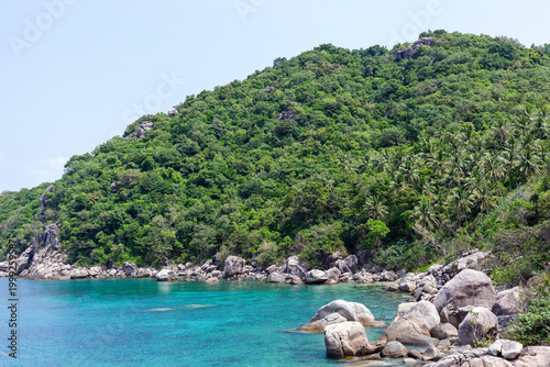 Mango Bay Koh Tao Thailand. Turquoise water meets rocky shoreline. Lush green forest covers the steep hills. Tropical island landscape under clear sky.