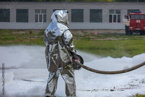 Firefighters use a foam generator to supply foam to extinguish the fire. Firefighters in protective clothing against high temperatures in a fire. Firefighters in shiny suits with foam generators.