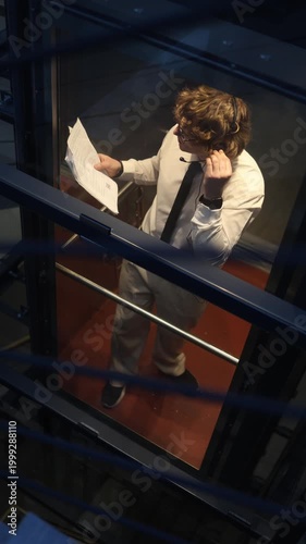 Vertical screen businessman works late in elevator talking with client on headset and reviewing documents showing dedication overtime effort and finishing tasks until the end
