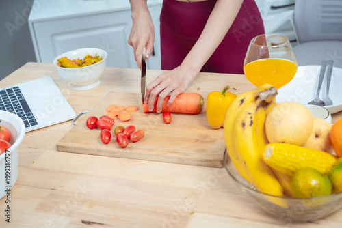 Young woman at kitchen table holding tomato and orange juice, surrounded by fresh vegetables and fruit, laptop nearby; healthy diet, nutrition and wellness concept in bright modern home.
