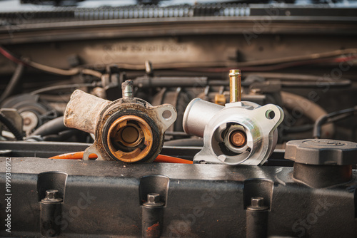 Comparison of an old, rusted car thermostat housing and a new replacement part on an engine, demonstrating automotive maintenance and repair concepts.