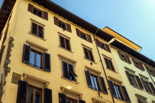 Facade Of Traditional Italian Historic Buildings In Florence Tuscany Italy With Ochre Yellow Stucco And Dark Green And Brown Wooden Window Shutters Under A Clear Blue Sky On A Sunny Day