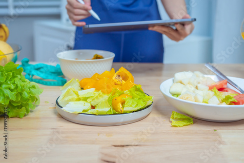 Young woman at kitchen table holding tomato and orange juice, surrounded by fresh vegetables and fruit, laptop nearby; healthy diet, nutrition and wellness concept in bright modern home.