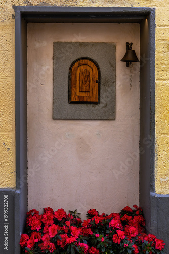 Charming Tuscan Style Architectural Niche And Shutter Inside An Old Building Facade With Arched Wooden Hatch A Hanging Brass Bell And Vibrant Red Azalea Flowers Inside The Recess At The Base