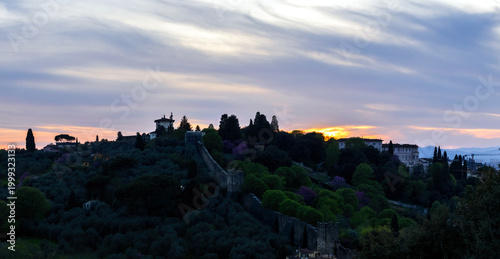 Panoramic Sunset View Of The Florentine Hills In Italy With Silhouetted Cypress Trees An Ancient City Wall And A Historical Villa Under A Cloudy Sky