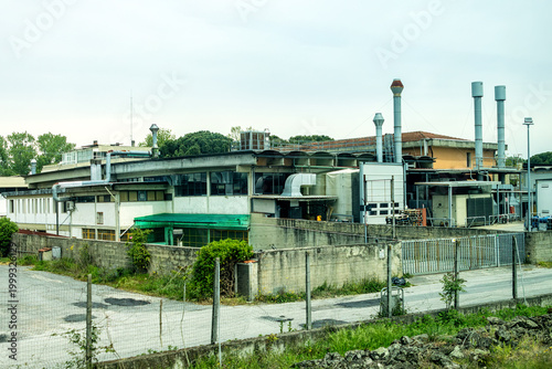 An Industrial Factory Complex with a Series of External Vents, Pipes, and HVAC Units Under an Overcast Sky with Concrete Walls and a Metal Fence in the Foreground