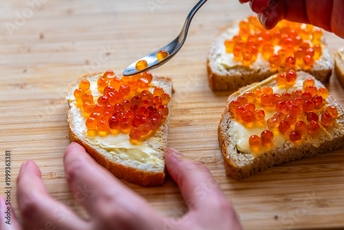 Bread butter sandwich with caviar, woman hand spreading fish roe with spoon macro closeup of wooden cutting board at home kitchen