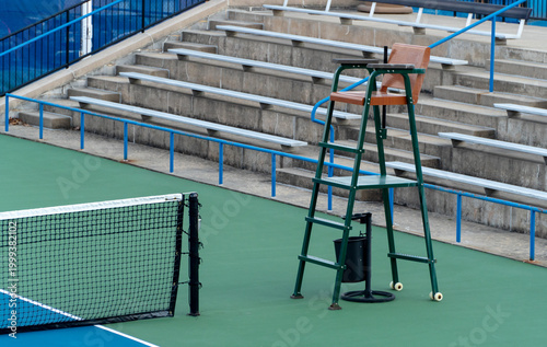 Foto Empty outdoor tennis court featuring a prominent green umpire chair, black net, and concrete bleachers with blue railings in the background, ready for play or a championship match