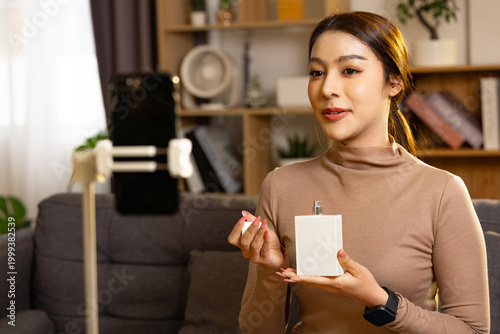 Happy young woman presenting perfume bottle to smartphone camera on tripod while recording video vlog at home in cozy living room setting