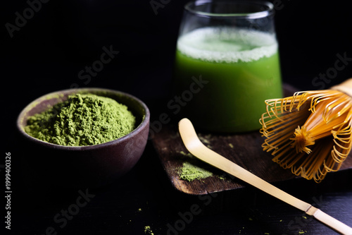 Green matcha drink with foam in a glass, styled with matcha powder in bowl, bamboo whisk and spoon.