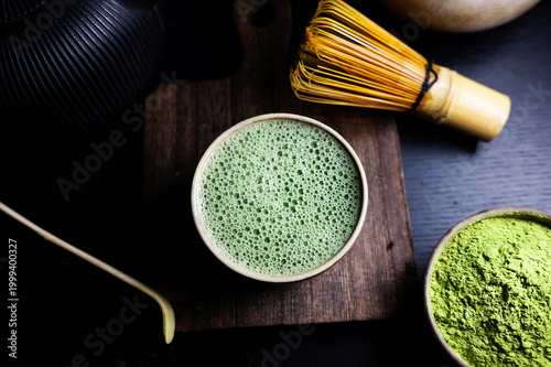 Freshly prepared matcha tea with rich foam in a cup, served with matcha powder, bamboo whisk and spoon.