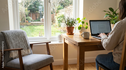 Cozy Workspace: A person immersed in work at a wooden table in a sunlit room, embraced by a sense of comfort and serenity. The soft glow illuminates indoor plants and the inviting backyard.