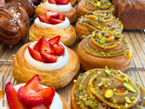 Danish pastries with strawberry cream and pistachio topping arranged closely on bakery rack. Sweet dessert selection, pastry variety, confectionery display and food styling concept.