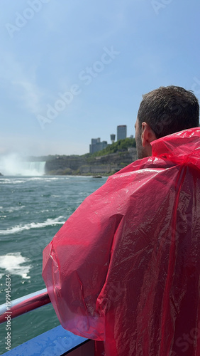 A tourist in a red poncho onboard a boat approaching Niagara Falls, with mist rising and lush cliffs under a sunny sky, Ontario, Canada.