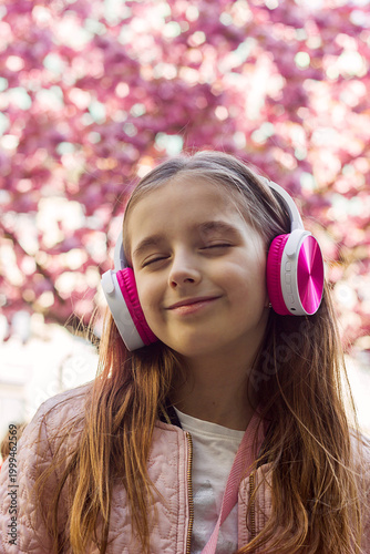 A beautiful little girl listens to music on headphones against the backdrop of flowering trees, close-up portrait