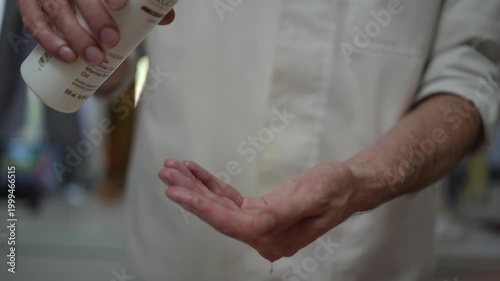 Unrecognizable male masseur pouring transparent massage oil from a white bottle onto his hands, preparing for a professional spa treatment
