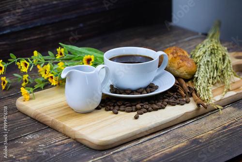 Still life coffee scene with cup of black coffee, bread, coffee beans, sunflower and cinnamon on wooden background, warm cozy cafe