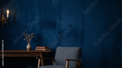 A cozy study room with a chair, table, vase, books, and sconce against a dark blue wall.