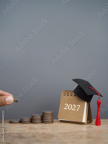 Hand putting coin into stack of coins and 2027 Desk calendar with graduation hat. The concept of saving money for education, student loan, scholarship, tuition fees in year 2027