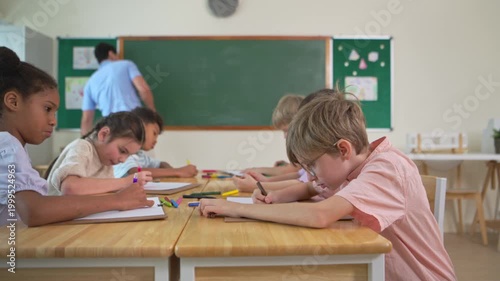 Diverse elementary students drawing and learning together in an international classroom while teacher writes on chalkboard