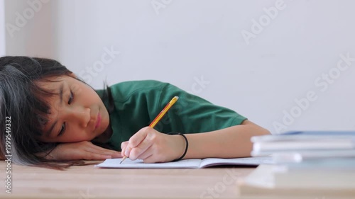 Close up selective focus of a tired Asian kid girl resting her head on the desk. Stressed and exhausted from studying too much homework in bright daylight. Education burnout and overwork concept.