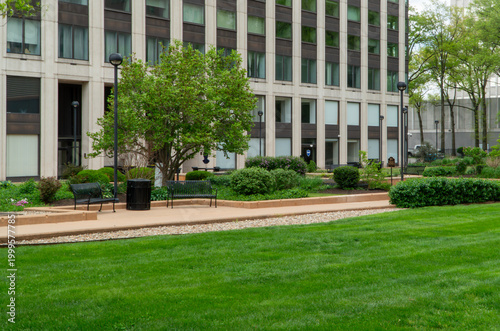 Vibrant green lawn in a peaceful urban park setting with modern office building facade, black benches, pathways, and diverse lush landscaping, ideal for corporate themes.