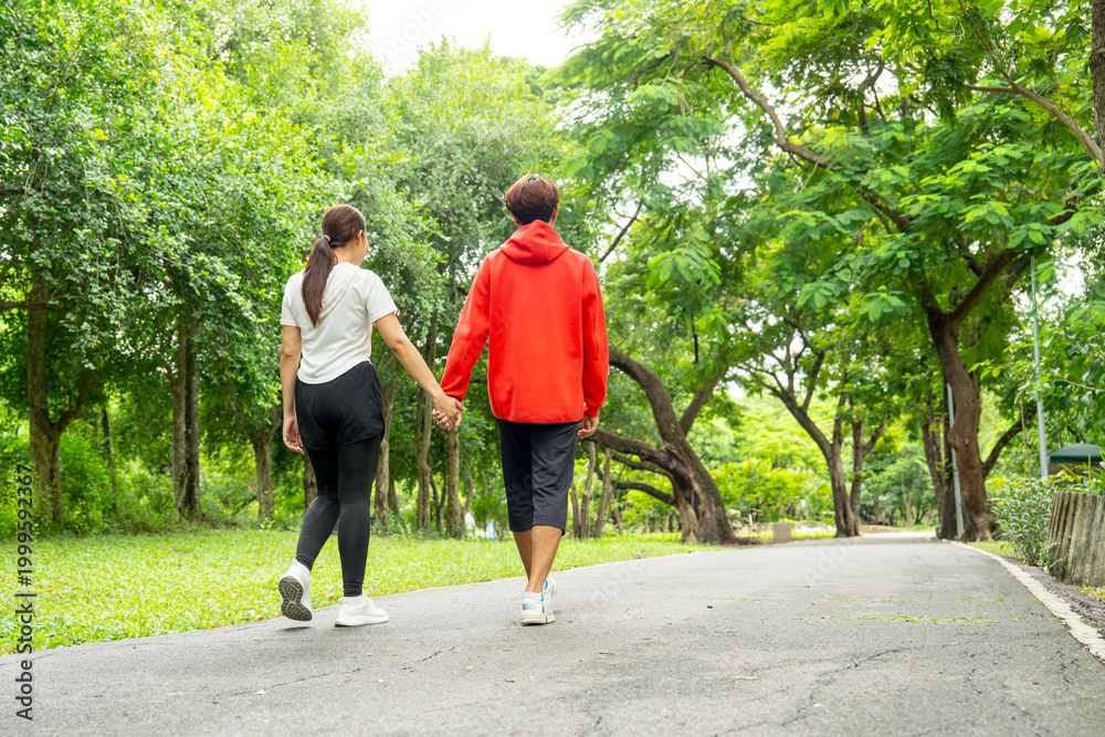 Fototapeta premium Back of Asian man and woman hold hands and walk together along the road in public park with day light for exercise and good activity for better relationship and healthy.
