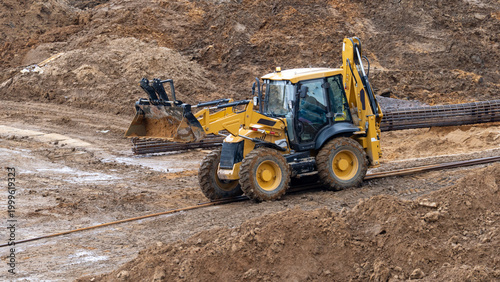 Yellow backhoe loader digs into muddy terrain. Its large tires sink into wet earth. The machine moves soil for land development. Construction site activity is clearly shown