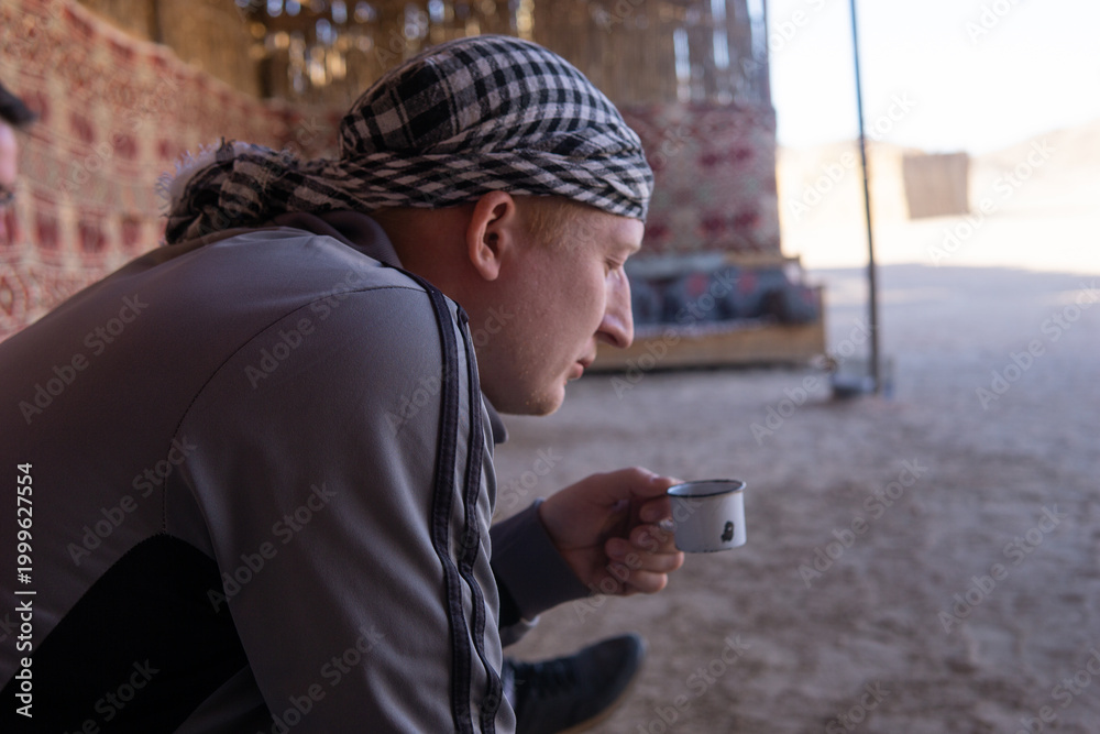 Fototapeta premium Man wearing a keffiyeh and grey jacket sits holding a small cup. Traveler enjoying a hot drink in a traditional bedouin tent.