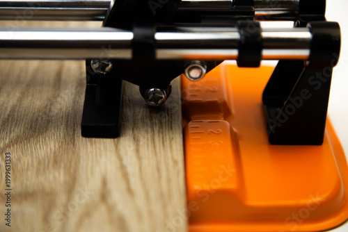 Close-up side view of an orange mechanical tile cutter, which fixed ceramic tiles of wooden colors, above it is a cutting disc on a white background. The concept of renovation, tiling, construction