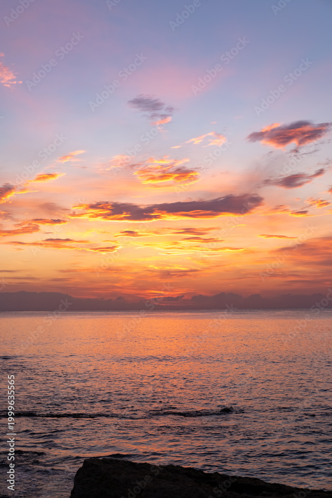Fototapeta premium Vibrant sunset over calm ocean waters with colorful clouds reflecting on the surface, rocky shoreline visible in the foreground and distant mountains on the horizon