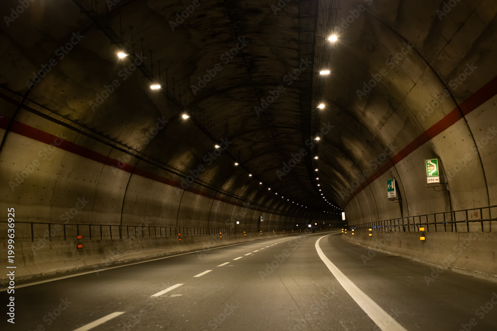 Fototapeta premium Dark concrete tunnel with illuminated lights and curved road, featuring road signs and safety barriers along the sides, creating a dramatic perspective view