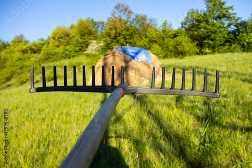Photos Close perspective of a hay rake in a green field leading toward a haystack, creating a dynamic composition that highlights farming tools and countryside agricultural work