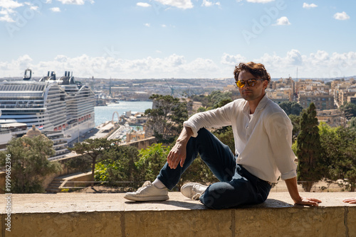 Young man in sunglasses sitting on a historic wall with a giant cruise ship in the background. Mediterranean travel concept in Valletta, Malta.