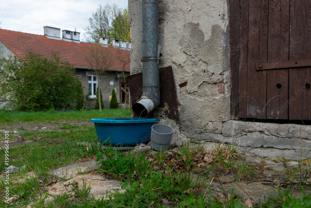 Obraz premium Old metal rain gutter on weathered wall draining into a blue plastic tub beside rustic buildings and grass. Useful for rainwater collection, rural yard, drainage system, old architecture and household