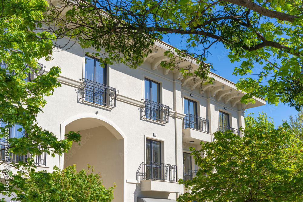 Fototapeta premium Classic-style light-colored building facade framed by vibrant green tree branches against a clear blue sky on a sunny day