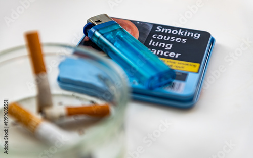 Photography An ashtray with cigarette ends and a lighter on a tobacco container with a health risk warning isolated on white