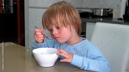 Boy with Down Syndrome in Blue Pajamas Eating Dinosaur Shaped Cereal with Milk in Kitchen
